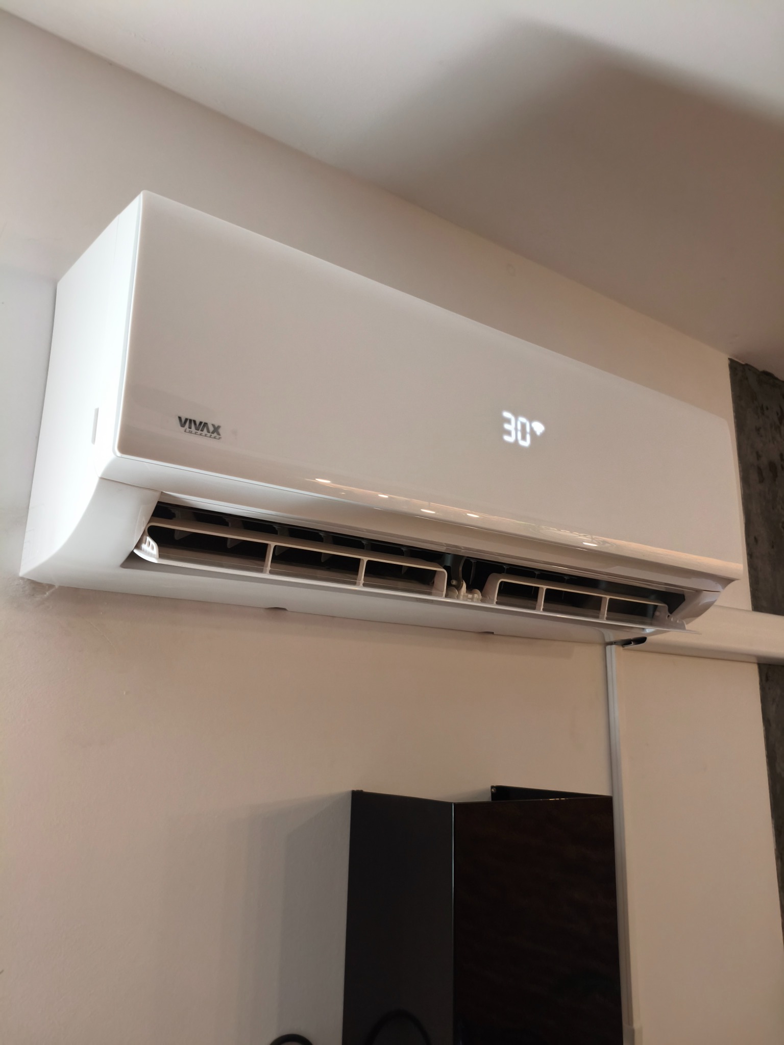 Biały klimatyzator Vivax z wyświetlaną temperaturą 30 stopni Celsjusza, zamontowany na jasnej ścianie w pomieszczeniu. Widoczny fragment szafki.