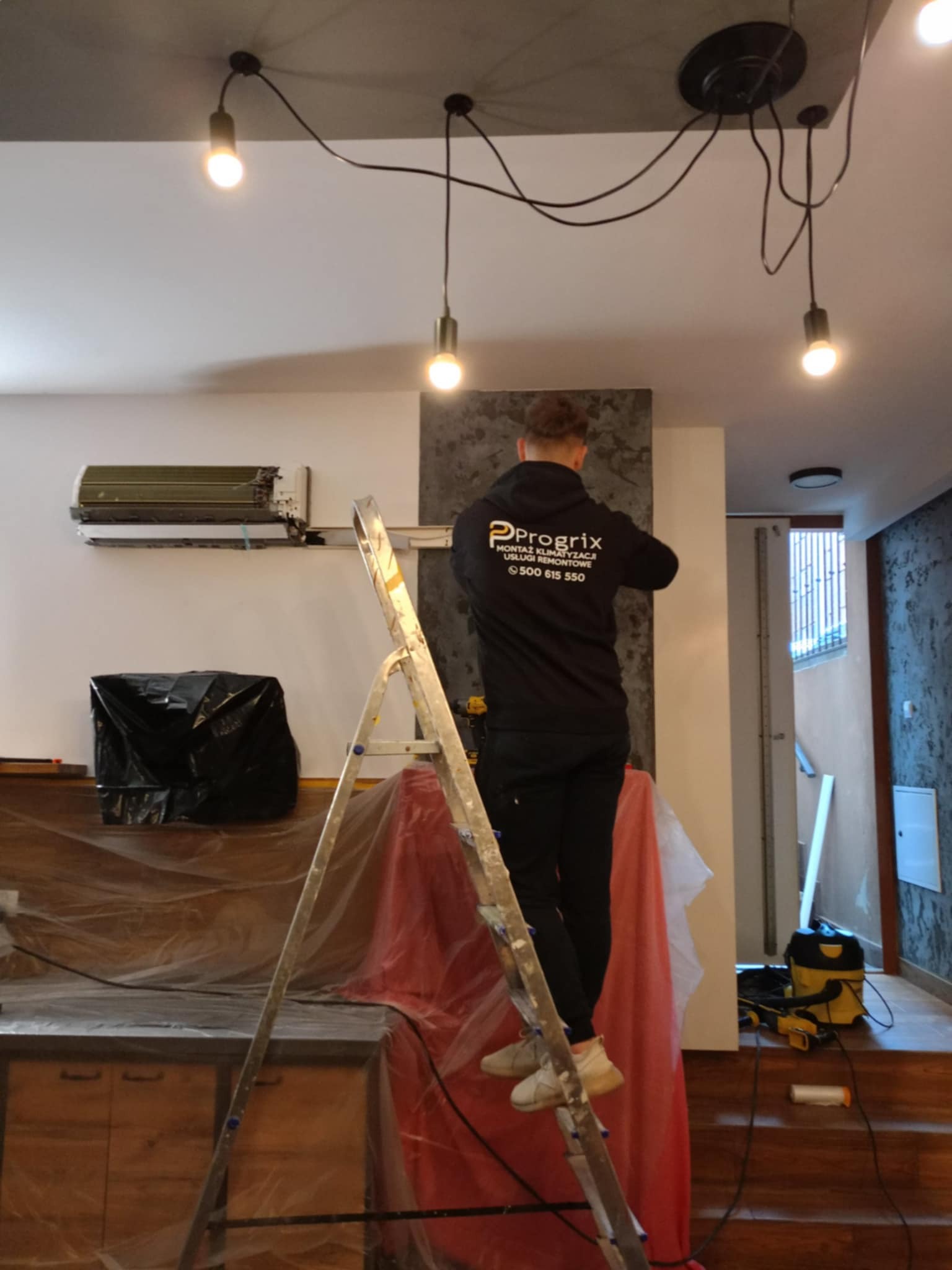 Mężczyzna na drabinie montuje klimatyzator. Widoczne narzędzia i zabezpieczone meble folią. Na bluzie logo firmy Progrrix.