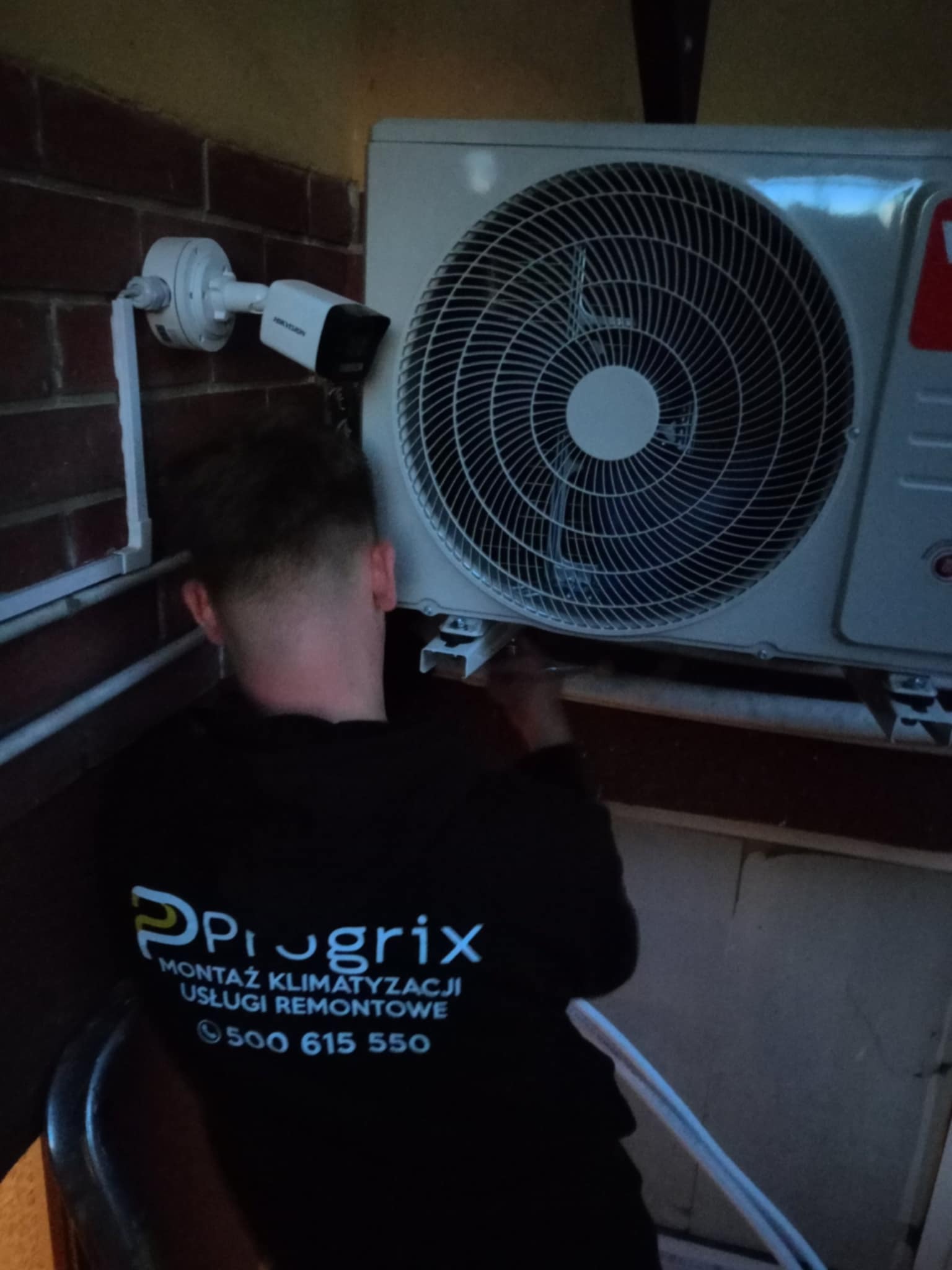 Montaż klimatyzacji zewnętrznej na ceglanej ścianie, widoczna kamera monitoringu i pracownik w firmowej bluzie z logo 'Prugrix' i numerem telefonu.