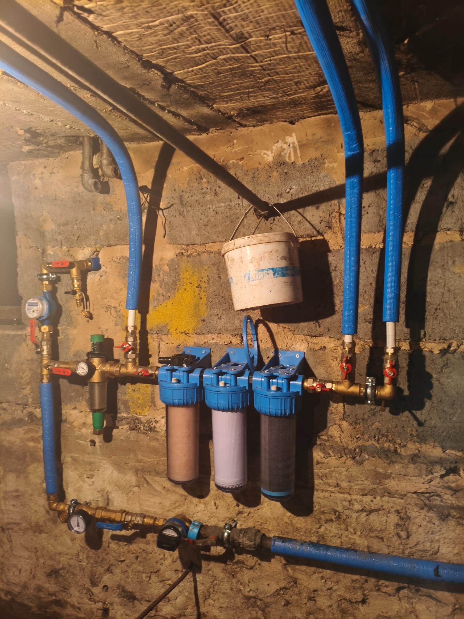 Instalacja filtrów wody z niebieskimi rurami na tle surowej ściany z cegły i wiszącym wiadrem. Widoczne zawory, manometry i liczniki.