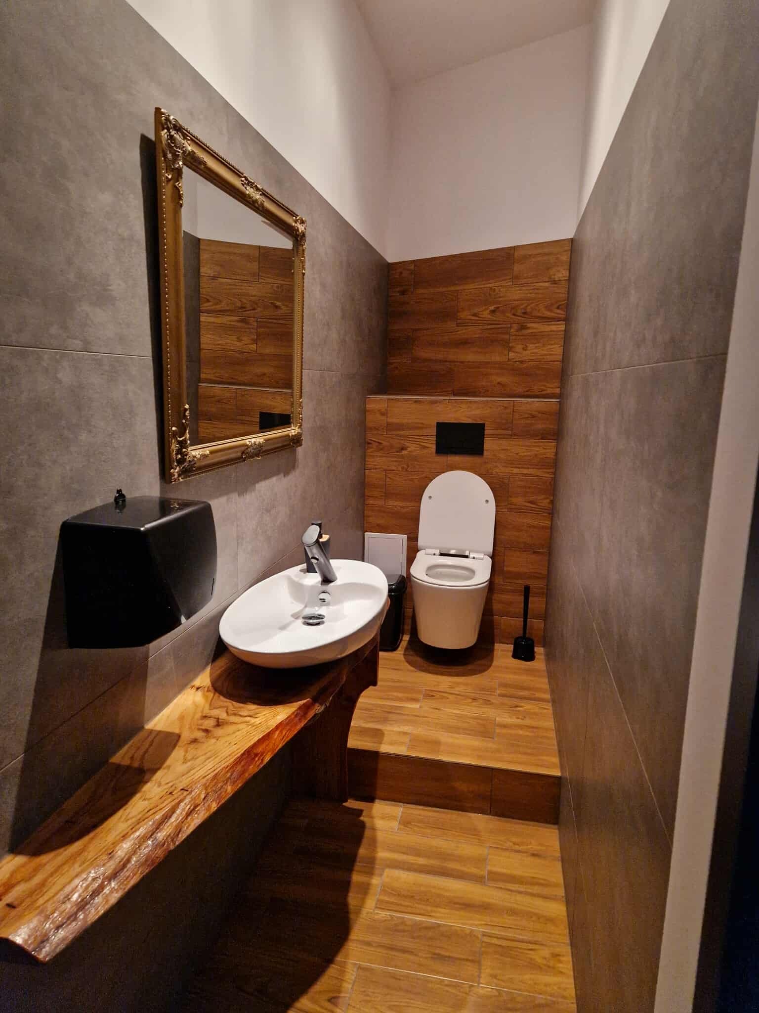 Nowoczesna łazienka z drewnianymi akcentami: blat pod umywalką z surowego drewna, lustro w złotej ramie i drewniana ściana za toaletą. Szare płytki na ścianach i podłodze.