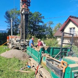 Precyzyjny Odwiert - Adrian Hołuj - Wiertnica do studni głębinowej w trakcie pracy na zewnątrz, z dwoma pracownikami i zielonym transportowym pojazdem gąsienicowym. W tle dom i błękitne niebo.