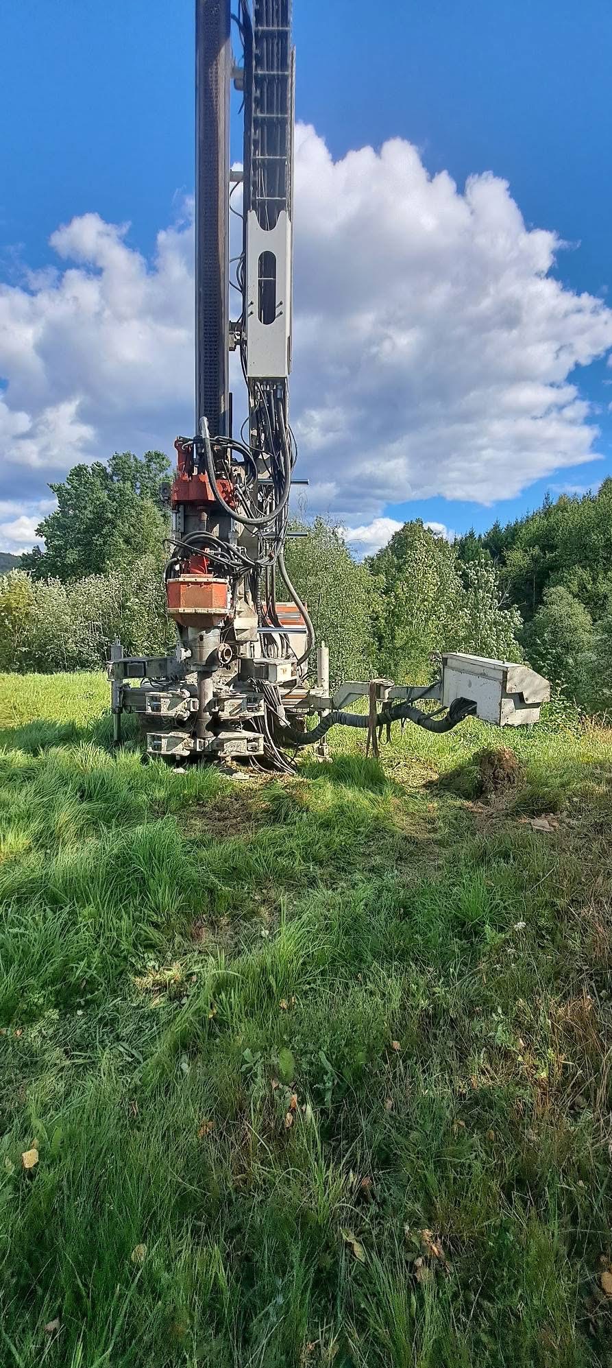 Wiertnica na trawiastym terenie, błękitne niebo z chmurami w tle. Widok na maszynę do wiercenia studni głębinowej w naturalnym otoczeniu.