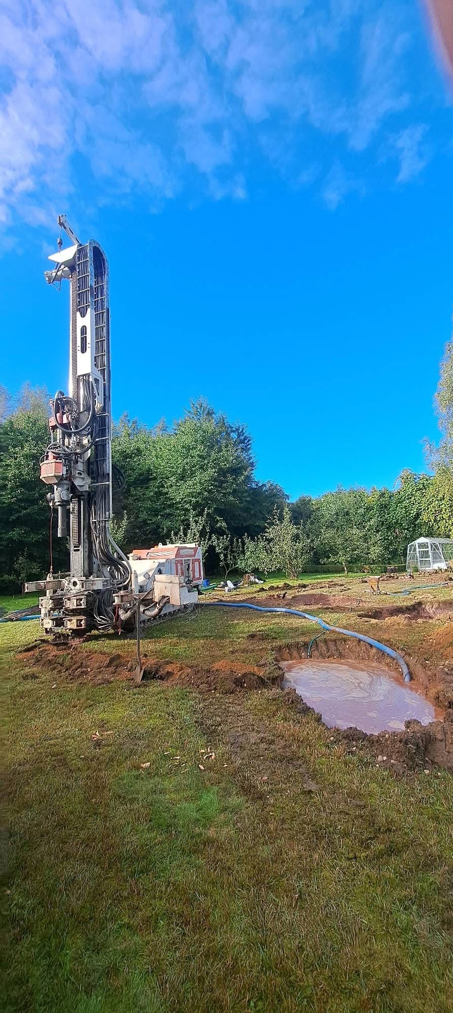 Wiertnica na trawniku, obok wykop z wodą. W tle drzewa i błękitne niebo. Prace ziemne związane z wierceniem studni.