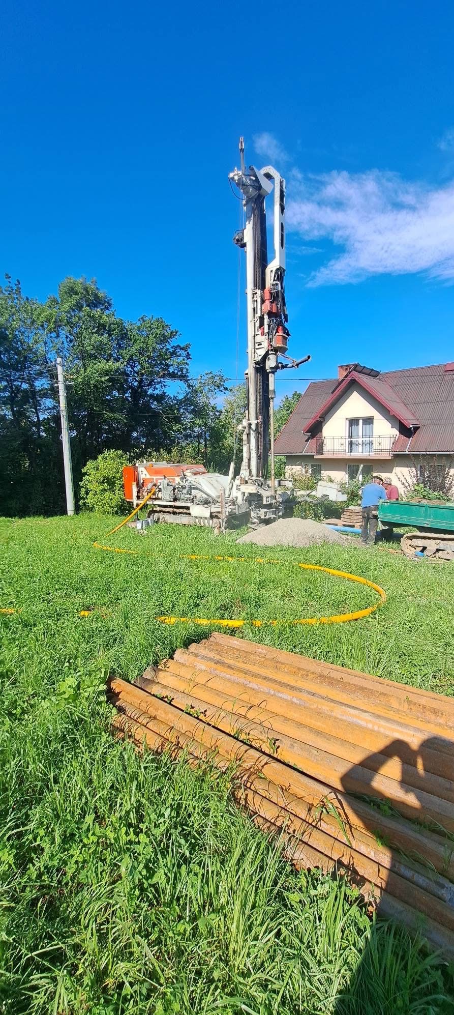 Wiertnica na trawniku przed domem podczas wiercenia studni. Żółte węże i rury w trawie. Błękitne niebo i zieleń drzew w tle. Działka z domem jednorodzinnym.