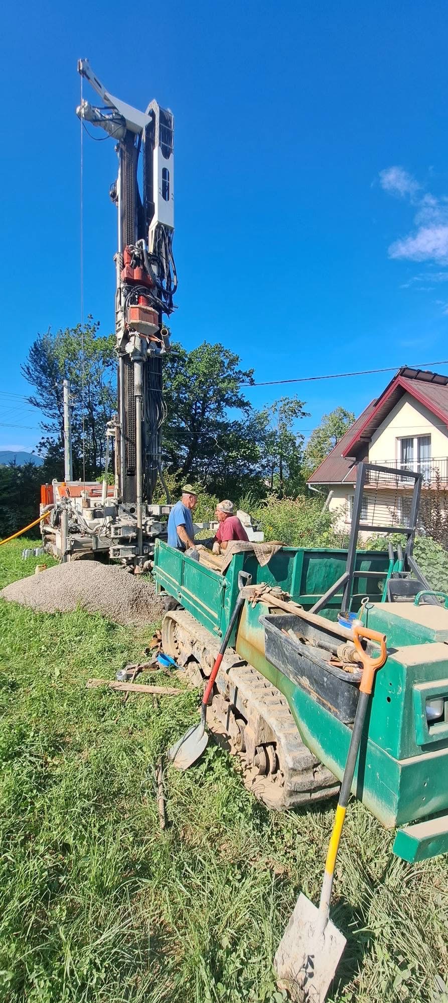 Wiertnica do studni głębinowej w trakcie pracy na zewnątrz, z dwoma pracownikami i zielonym transportowym pojazdem gąsienicowym. W tle dom i błękitne niebo.