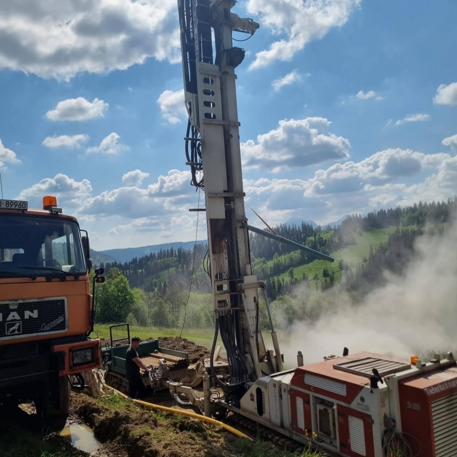 Wiercenie studni głębinowej w terenie górzystym. Widoczna maszyna wiertnicza, ciężarówka MAN i pracownik. W tle zielone wzgórza i błękitne niebo z chmurami.
