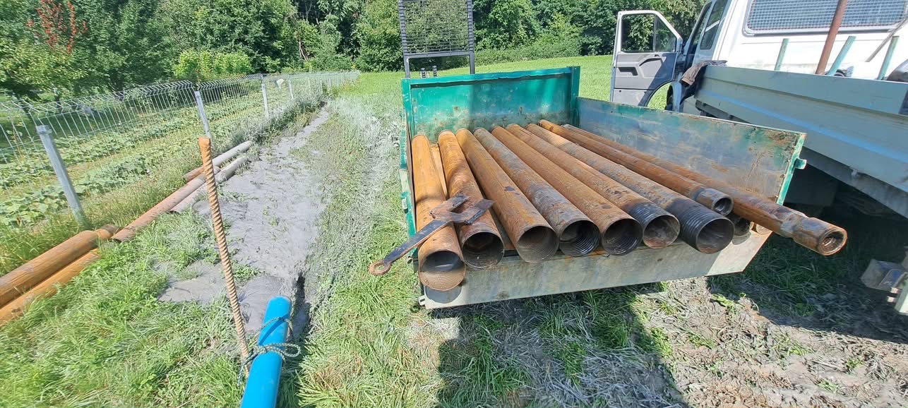 Rury do wiercenia studni głębinowych w skrzyni samochodu na tle zieleni i ogrodzenia. Widoczny sprzęt do łączenia rur.