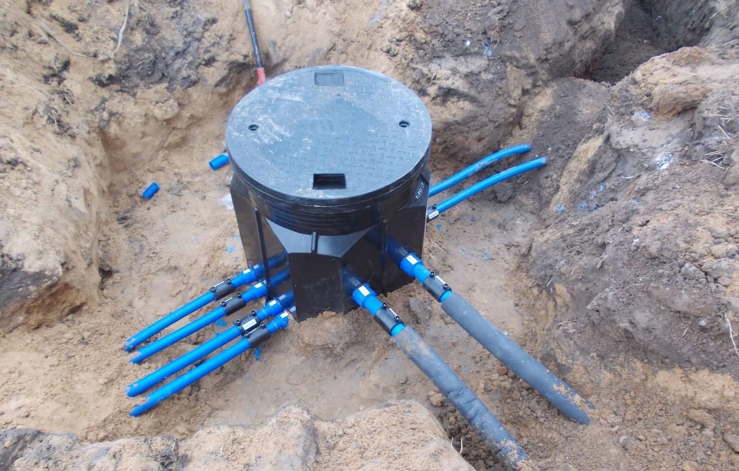 Czarna studzienka rewizyjna z niebieskimi rurami w wykopie ziemnym. Widoczne złącza i pokrywa studzienki. Ujęcie z góry, na ziemi widoczne ślady prac ziemnych.