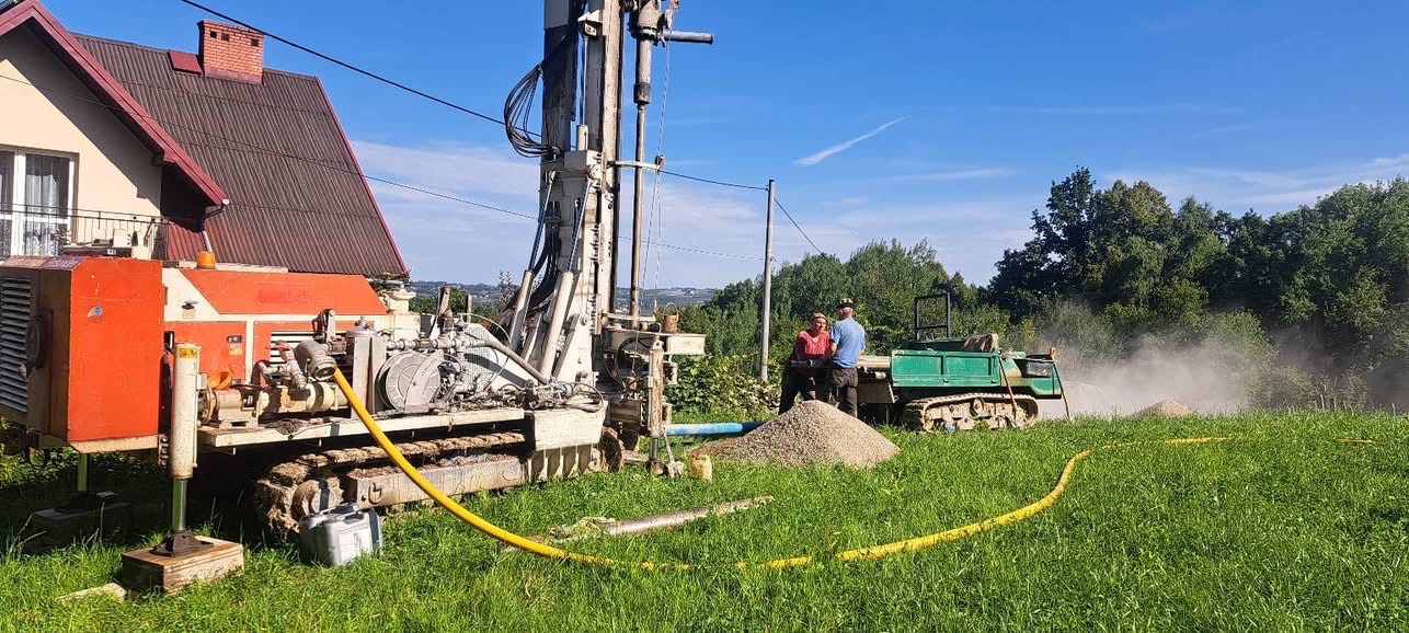 Wiertnica na gąsienicach podczas pracy przy domu. W tle dwójka pracowników i zielony transportowy pojazd gąsienicowy. Wokół zielona trawa i drzewa.