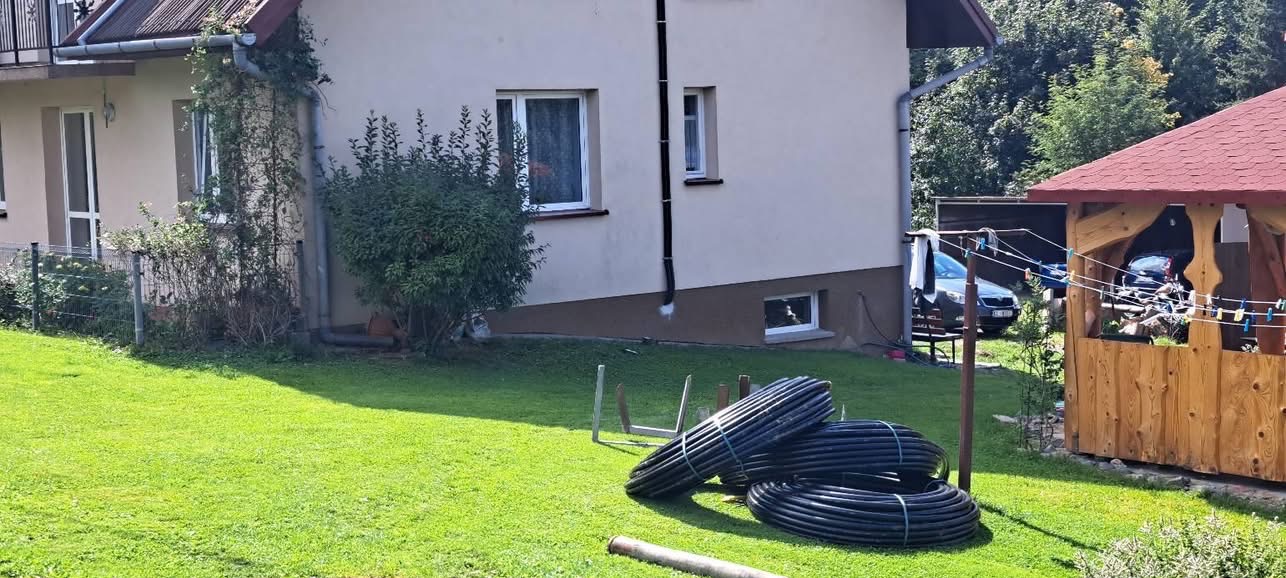 Zrolowane czarne rury na trawniku przed domem z beżową elewacją. Widoczny fragment ogrodu z altaną i suszarką na pranie. Przygotowanie do instalacji.