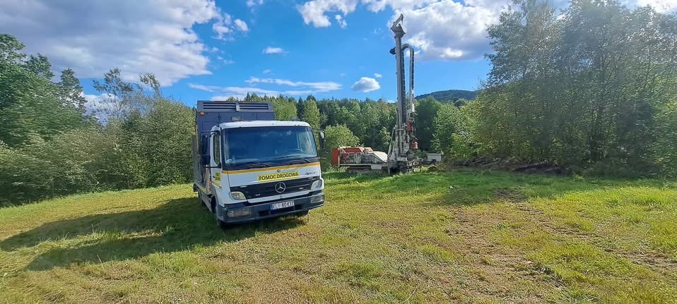 Wiertnica do studni głębinowych na zielonym terenie, obok zaparkowany samochód serwisowy z napisem 'Pomoc Drogowa'. Błękitne niebo z chmurami w tle.