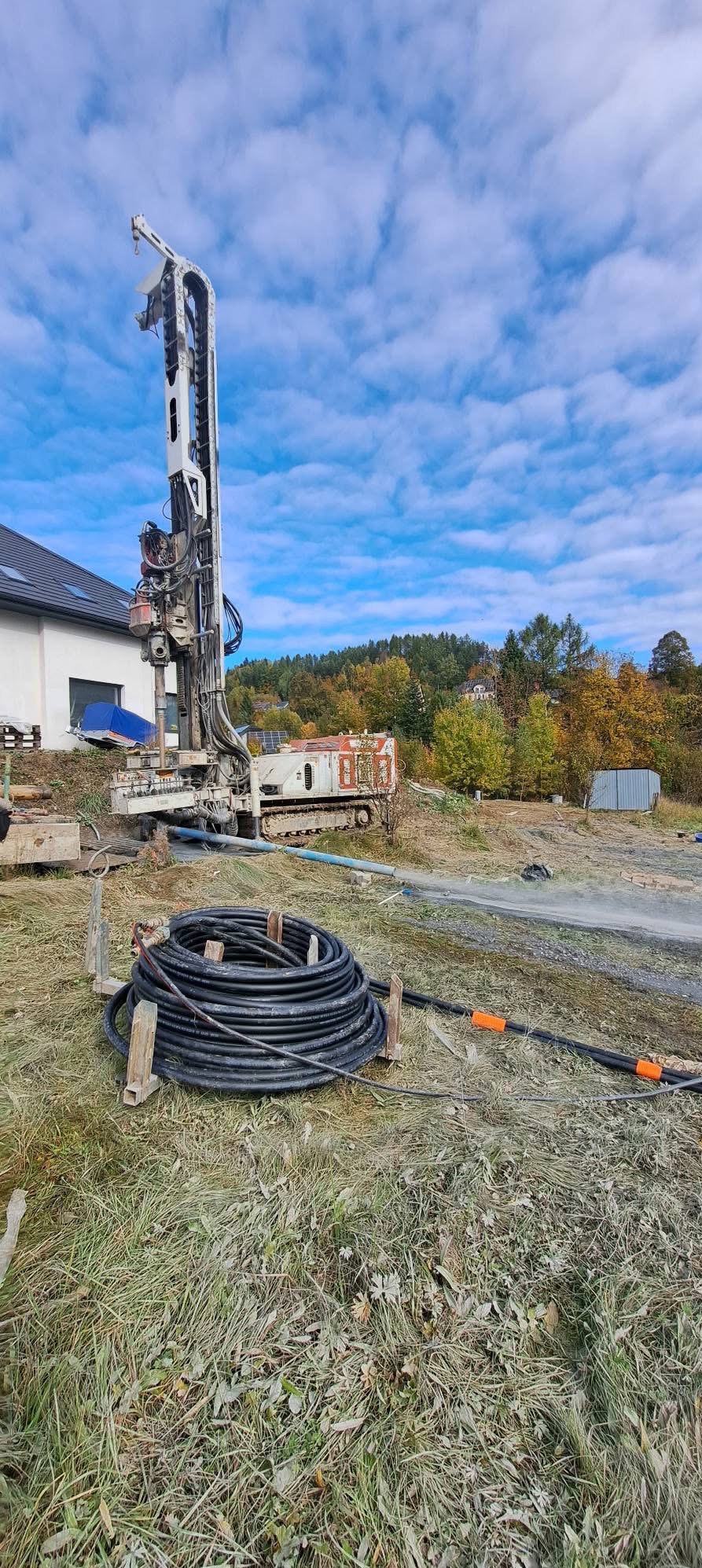 Wiertnica na gąsienicach podczas pracy przy domu jednorodzinnym. Na pierwszym planie zwój czarnych węży. Błękitne niebo z chmurami w tle.