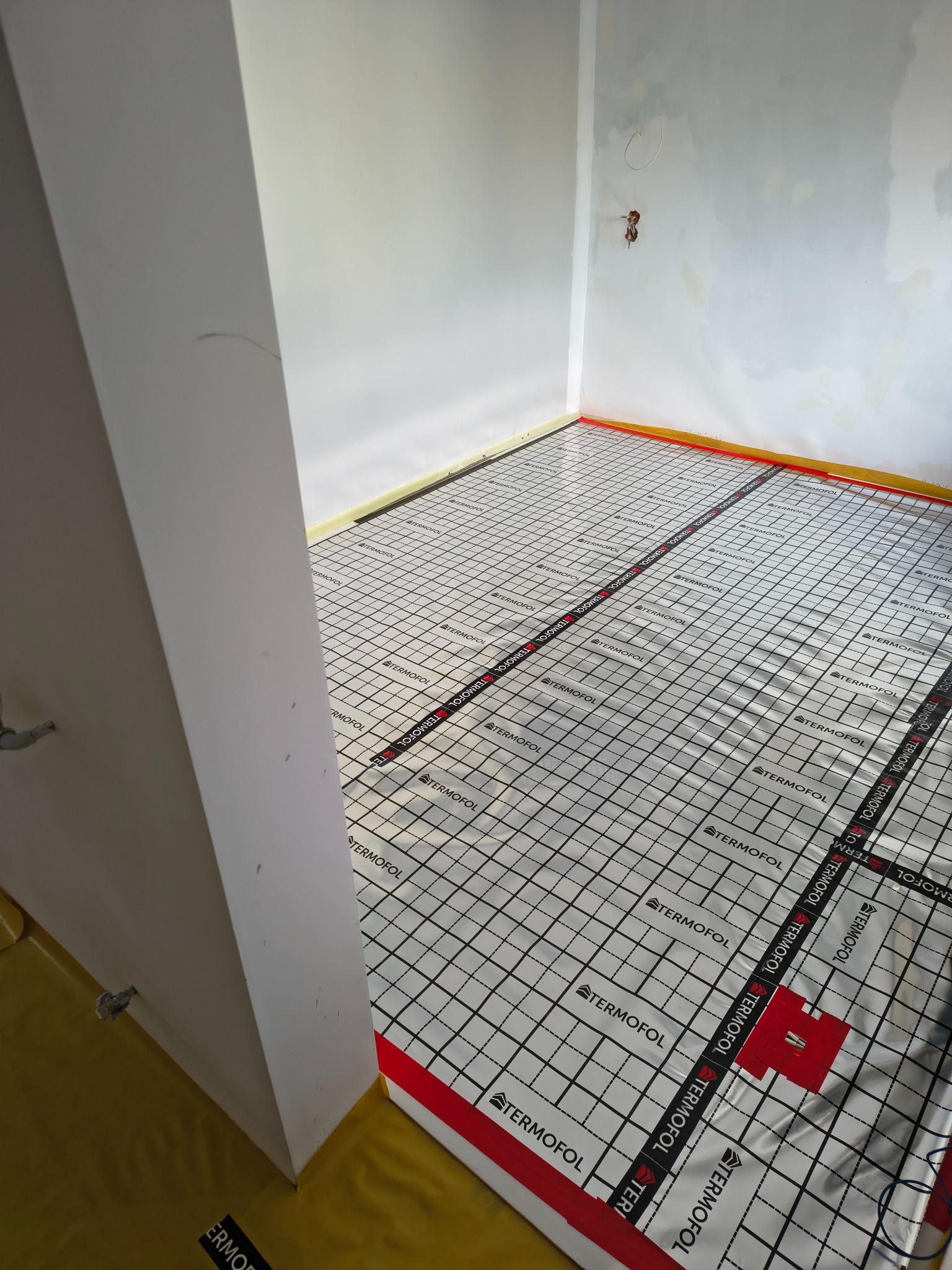 Montaż maty grzewczej Termofol na podłodze w nowym pomieszczeniu, przygotowanej do dalszych prac wykończeniowych. Widoczne taśmy mocujące i folia izolacyjna.