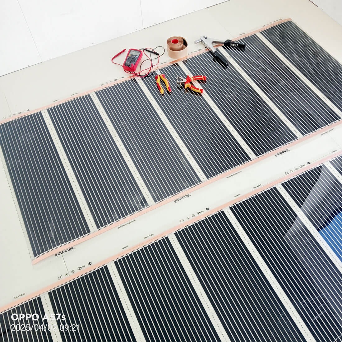 Rozłożone maty grzewcze na podłodze, widoczne narzędzia: miernik, taśma, szczypce. Czarno-białe pasy mat grzewczych ułożone równolegle. Widok z góry, jasne tło.