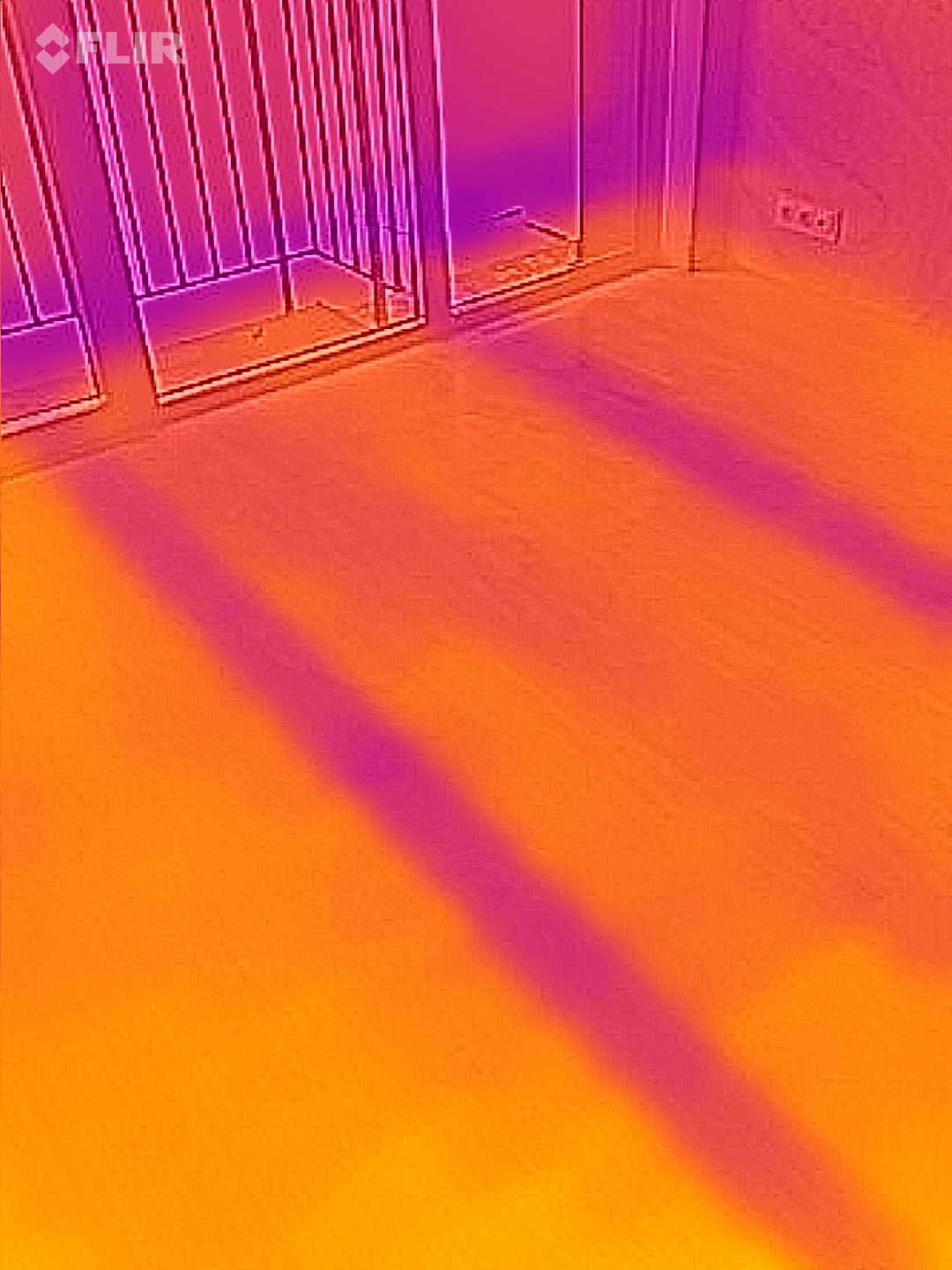 Termowizja podłogi z widocznymi przewodami grzewczymi, ukazująca rozkład temperatury. Widoczne okno balkonowe i gniazdka elektryczne na ścianie.