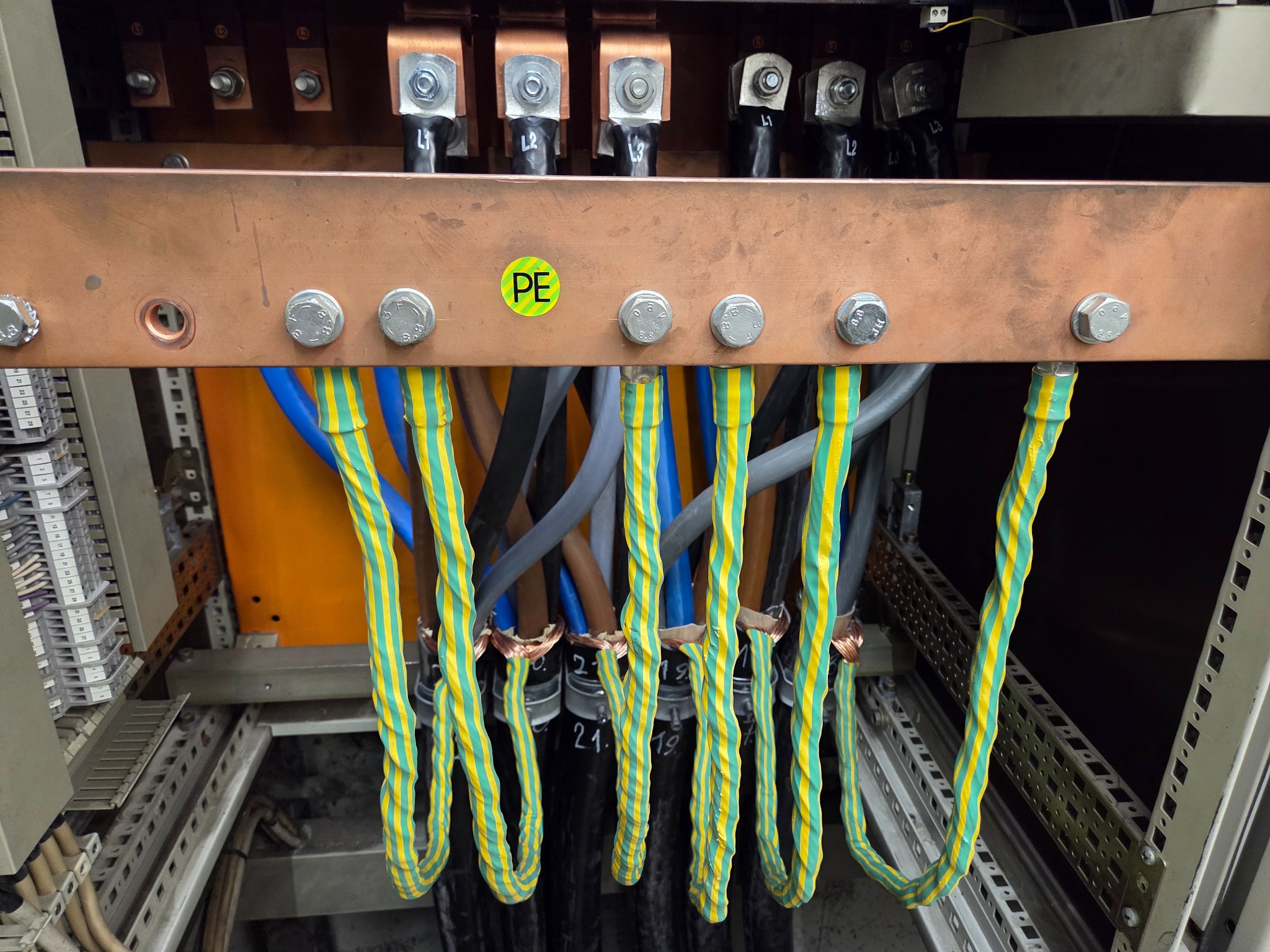 Szyna PE z podłączonymi przewodami w izolacji żółto-zielonej, niebieskiej i szarej. Widoczne śruby mocujące i oznaczenie PE na zielonej naklejce.