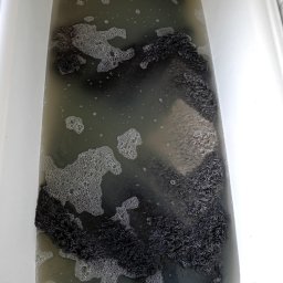 Shinycar24h - Czarny dywan shaggy w wannie wypełnionej brudną wodą z pianą, ukazujący proces prania dywanu w domowych warunkach. Widok z góry na wannę.