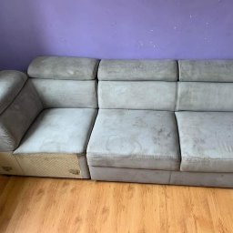 Shinycar24h - Szara, modułowa kanapa z widocznymi plamami na tapicerce, ustawiona na drewnianej podłodze przed fioletową ścianą. Widoczne detale konstrukcyjne.