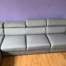 Shinycar24h - Szara, modułowa kanapa z regulowanymi zagłówkami na tle fioletowej ściany i drewnianej podłogi. Widoczne ślady użytkowania na podłodze.