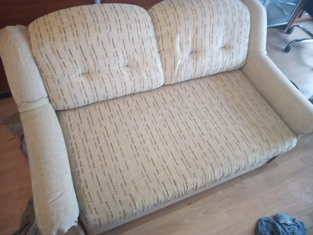 Dwuosobowa kanapa z beżową tapicerką w drobne, ciemne prążki, widoczna z góry pod kątem. Na podłodze leży szmatka.