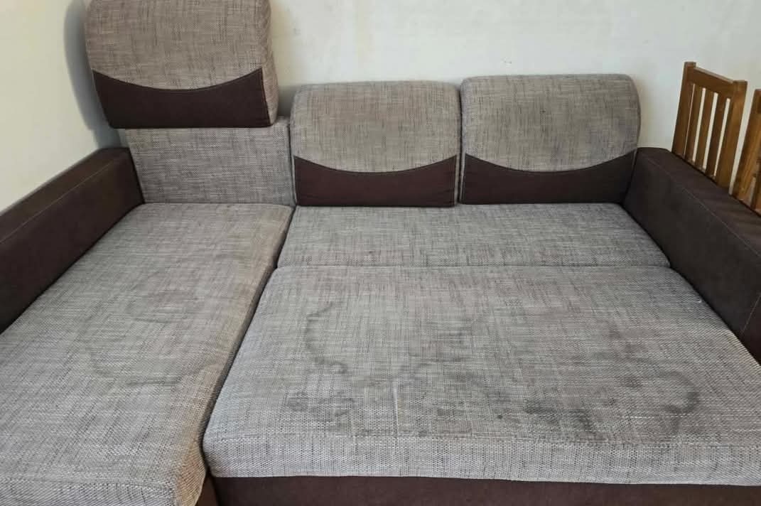 Szaro-brązowy narożnik z widocznymi plamami na siedzisku i oparciach, ukazujący potrzebę odświeżenia tapicerki. Widoczne drewniane elementy krzesła w tle.