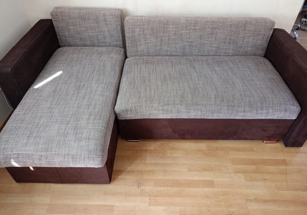 Rozłożysta, brązowo-szara kanapa narożna z tkaniny, ustawiona na drewnianej podłodze. Widoczne metalowe nóżki i subtelne światło padające na tapicerkę.