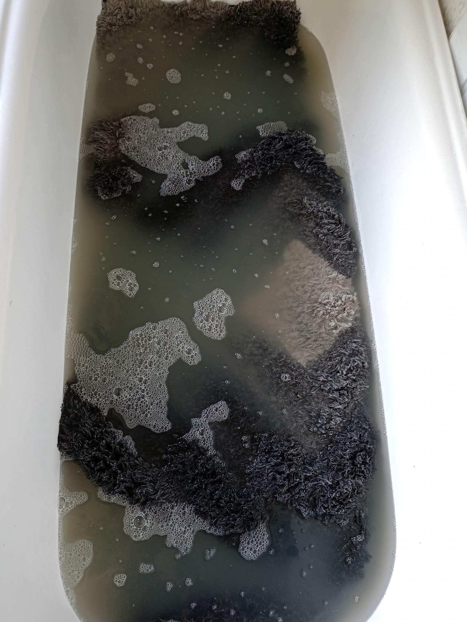 Czarny dywan shaggy w wannie wypełnionej brudną wodą z pianą, ukazujący proces prania dywanu w domowych warunkach. Widok z góry na wannę.