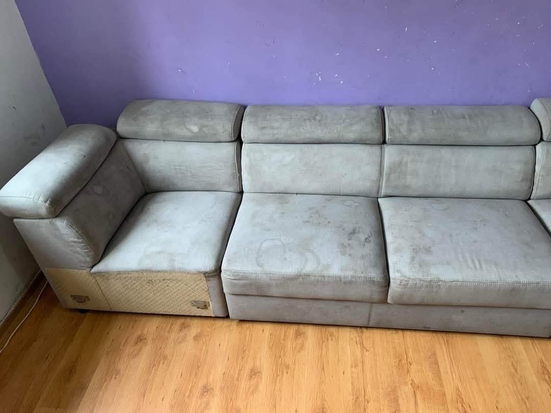 Szara, modułowa kanapa z widocznymi plamami na tapicerce, ustawiona na drewnianej podłodze przed fioletową ścianą. Widoczne detale konstrukcyjne.