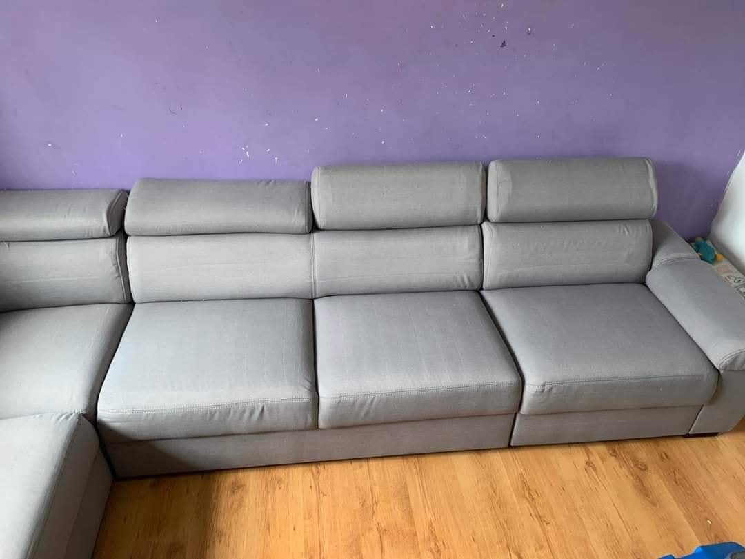 Szara, modułowa kanapa z regulowanymi zagłówkami na tle fioletowej ściany i drewnianej podłogi. Widoczne ślady użytkowania na podłodze.