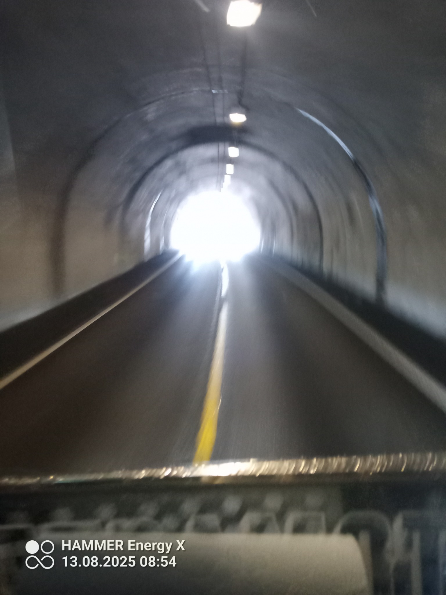 Wnętrze tunelu drogowego widziane z kabiny pojazdu. Perspektywa zza kierownicy, światło na końcu tunelu, żółta linia na jezdni. Ujęcie w ruchu, z widocznym pulpitem.