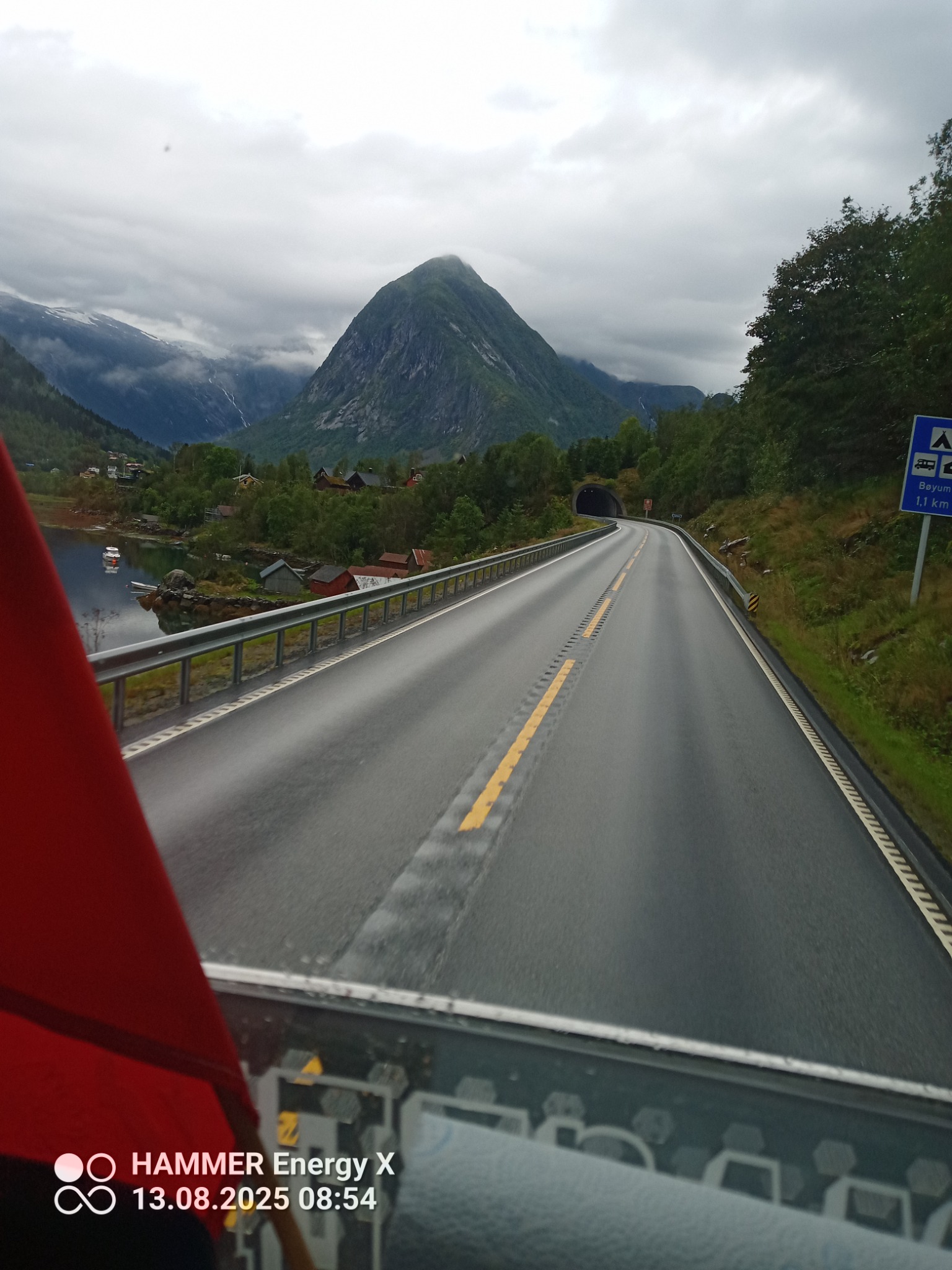 Widok z kabiny pojazdu na asfaltową drogę prowadzącą przez górzysty krajobraz Norwegii, z tunelem w oddali i jeziorem po lewej stronie. Pochmurne niebo.
