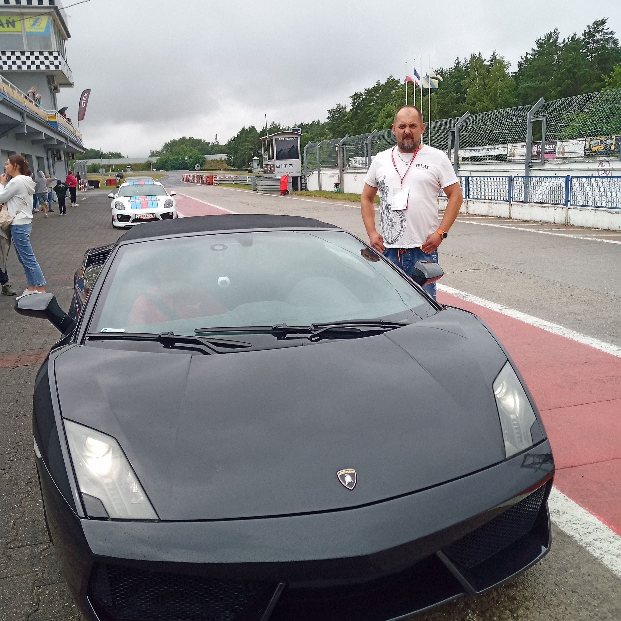 Czarny Lamborghini Gallardo Spyder na torze wyścigowym, z mężczyzną w tle i innymi autami. Pochmurne niebo, widoczny budynek kontrolny i flagi. Detal: logo Lamborghini na masce.