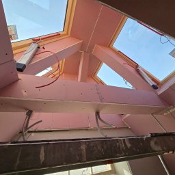 Krisputz - Montaż płyt gipsowo-kartonowych na poddaszu z oknami dachowymi. Widoczne drewniane elementy konstrukcyjne i instalacje elektryczne. Ujęcie z dołu, z drabiny.