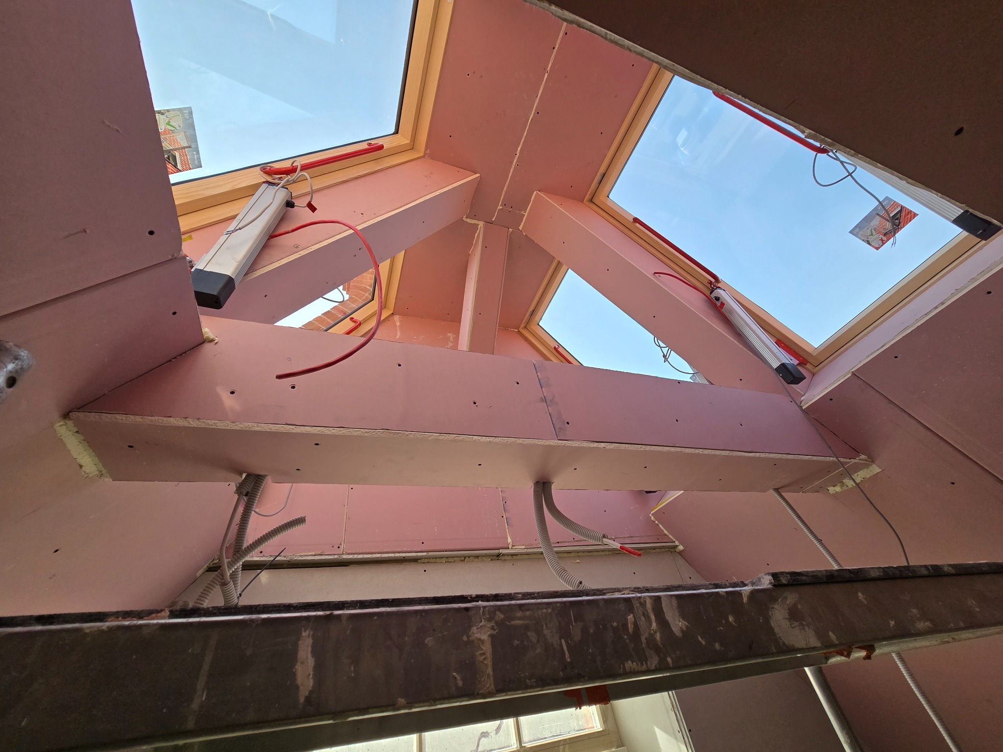 Montaż płyt gipsowo-kartonowych na poddaszu z oknami dachowymi. Widoczne drewniane elementy konstrukcyjne i instalacje elektryczne. Ujęcie z dołu, z drabiny.