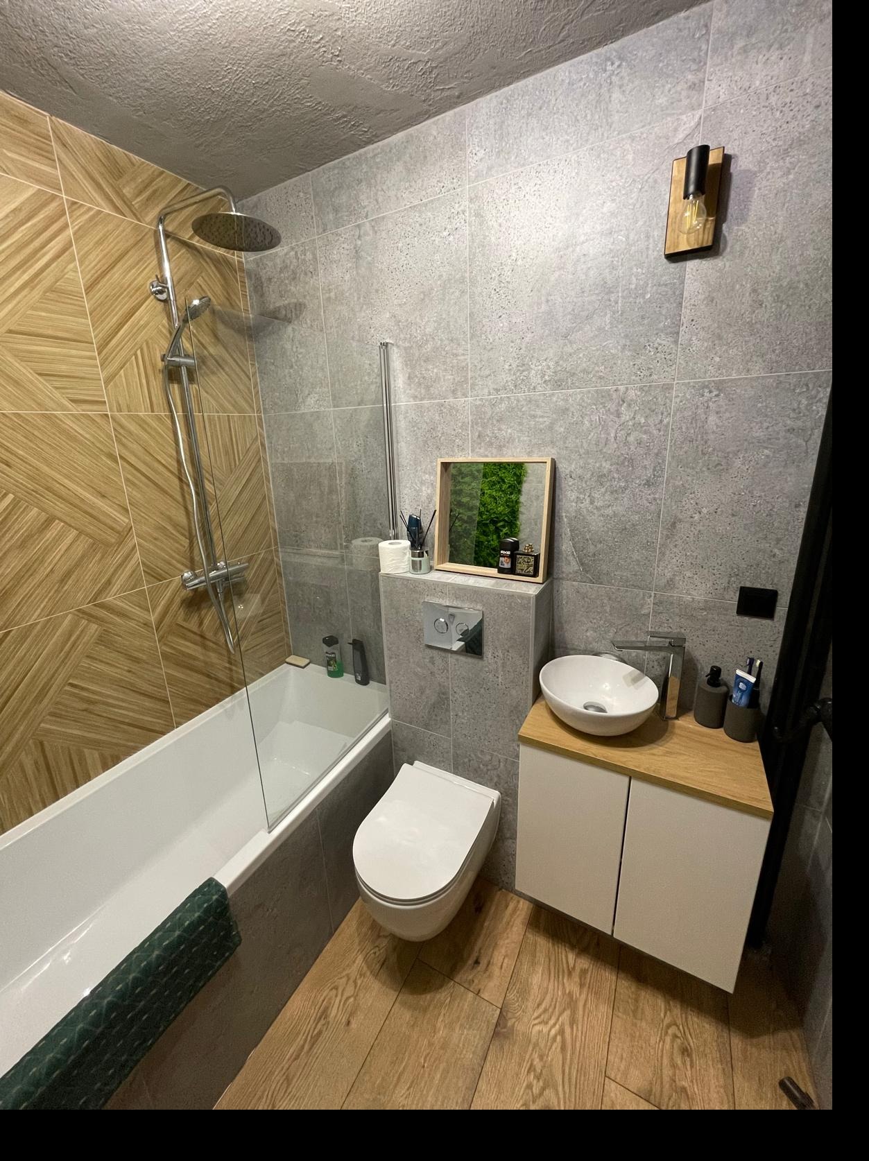 Nowoczesna łazienka po remoncie: drewnopodobne płytki, szare ściany, biała armatura, umywalka nablatowa i dekoracja z mchu w ramce. Oświetlenie punktowe.