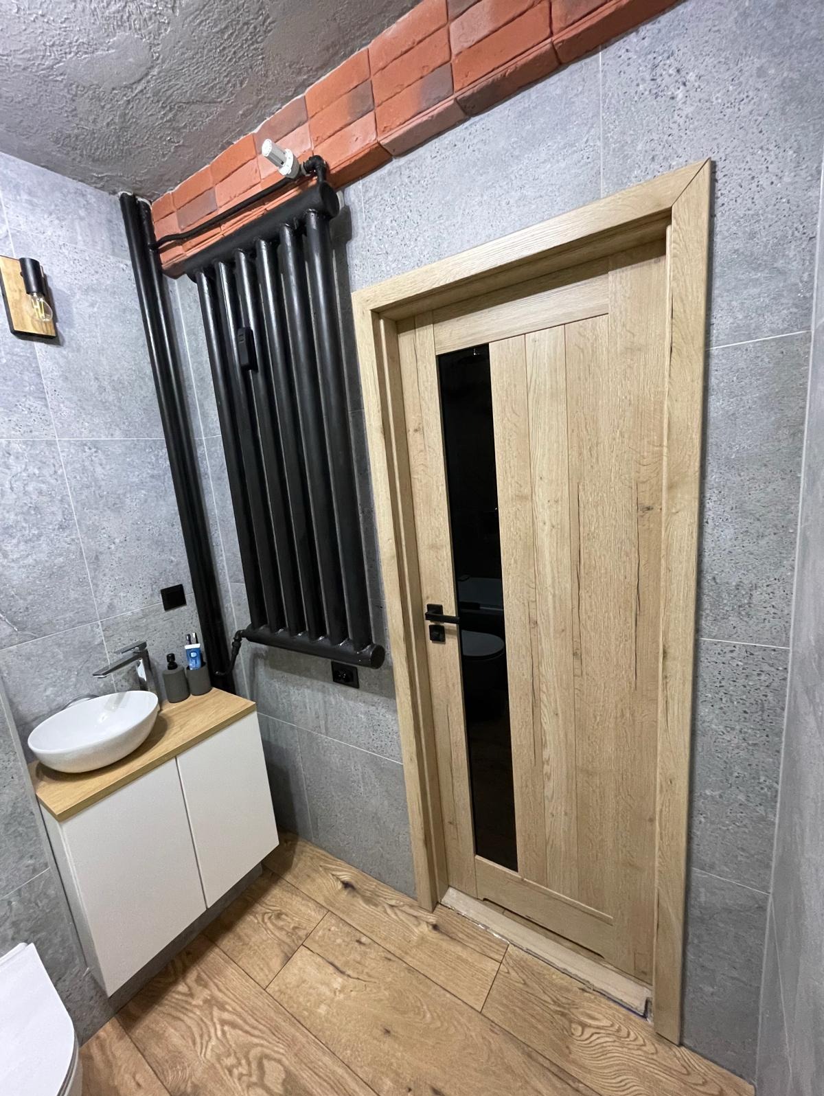 Nowoczesna łazienka z drewnianą podłogą i drzwiami z czarnym szkłem. Na ścianie szary gres, czarny grzejnik i umywalka nablatowa na białej szafce.