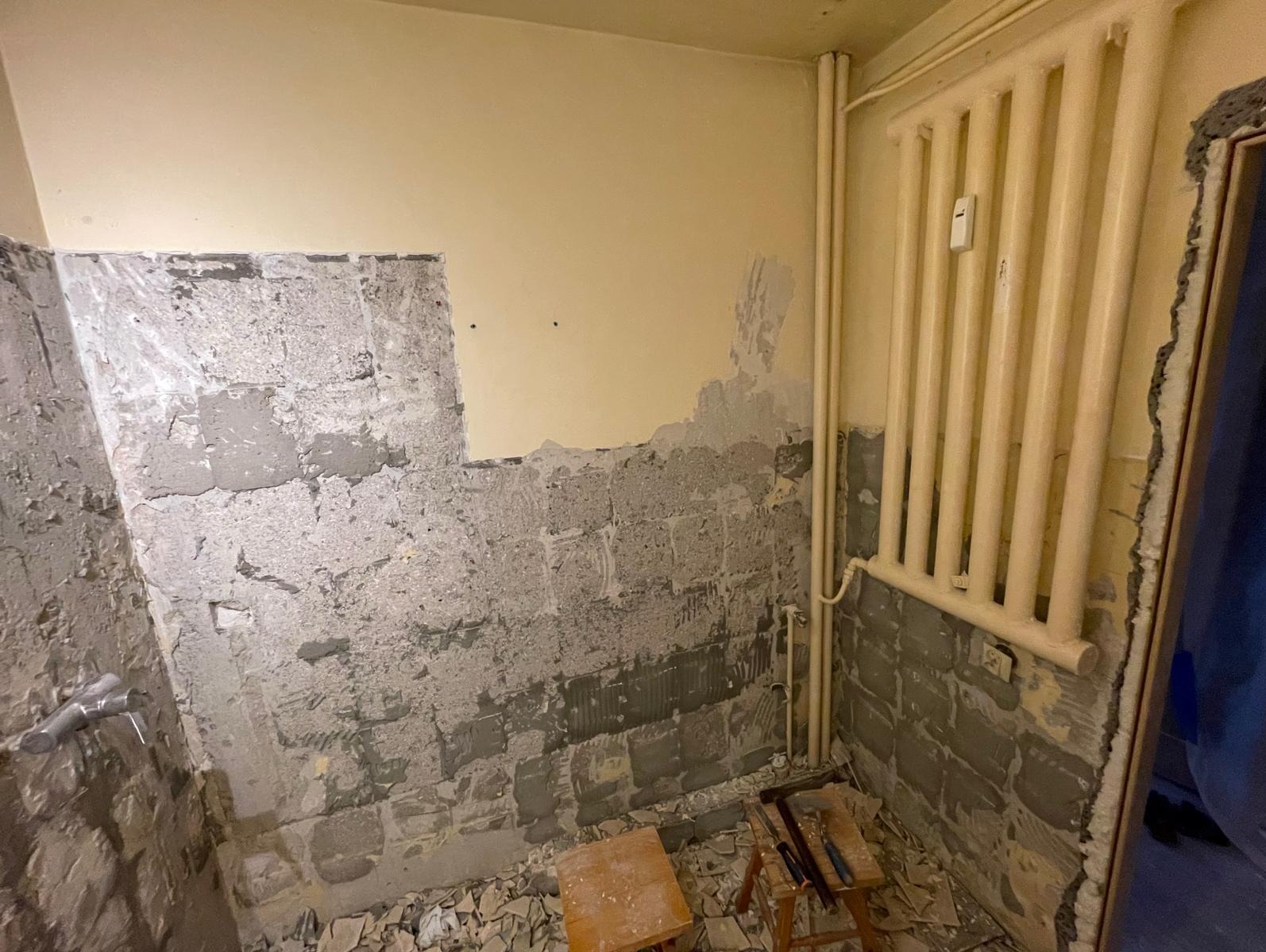 Demontaż płytek w łazience, ściany z resztkami kleju i gruzu, widoczny grzejnik i narzędzia glazurnicze. Etap przygotowania do remontu, surowe wykończenie.