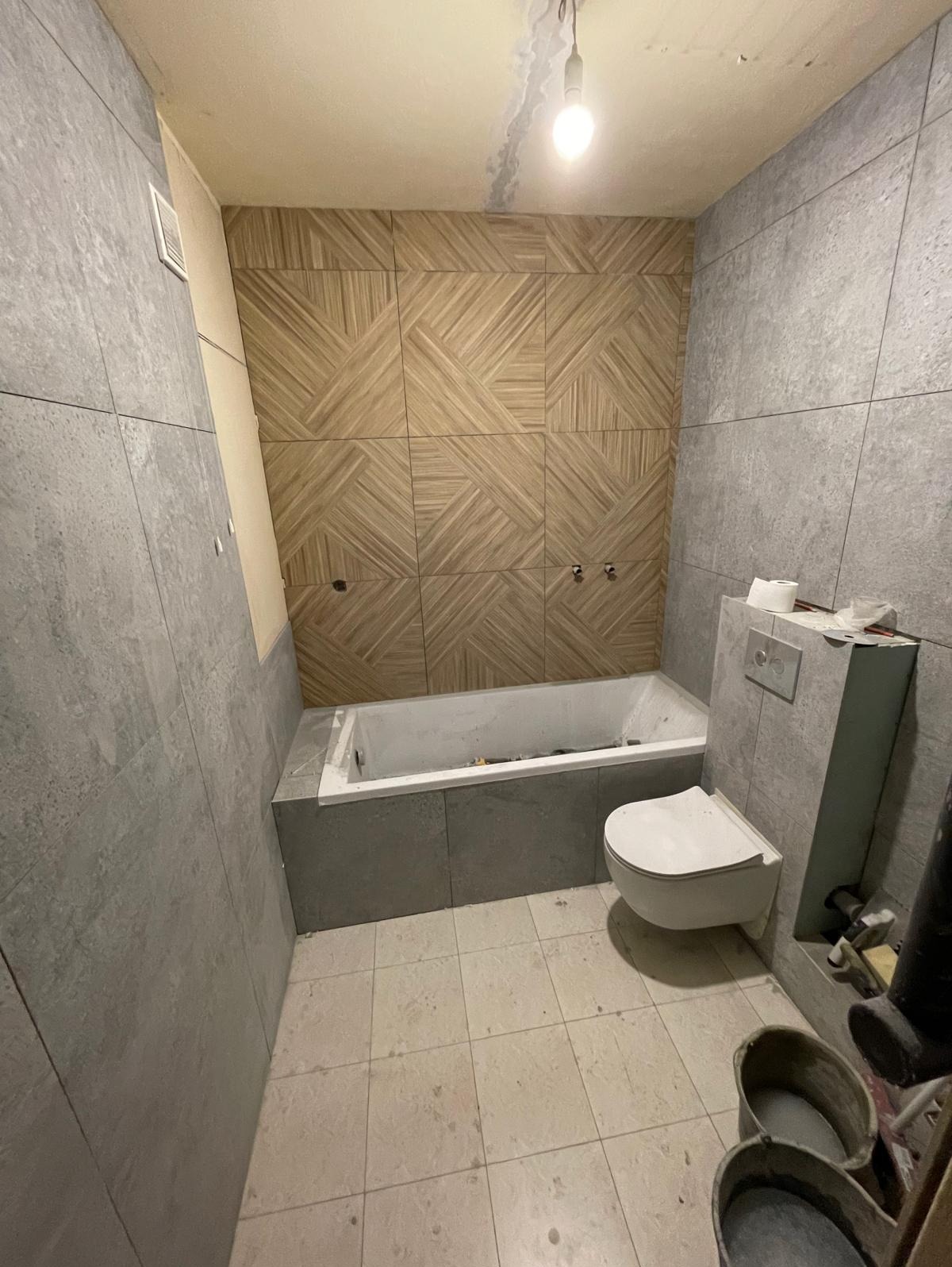 Wykończona łazienka z szarą i drewnopodobną glazurą ułożoną w geometryczny wzór na ścianie nad wanną. Widoczna toaleta i wiaderka z zaprawą.