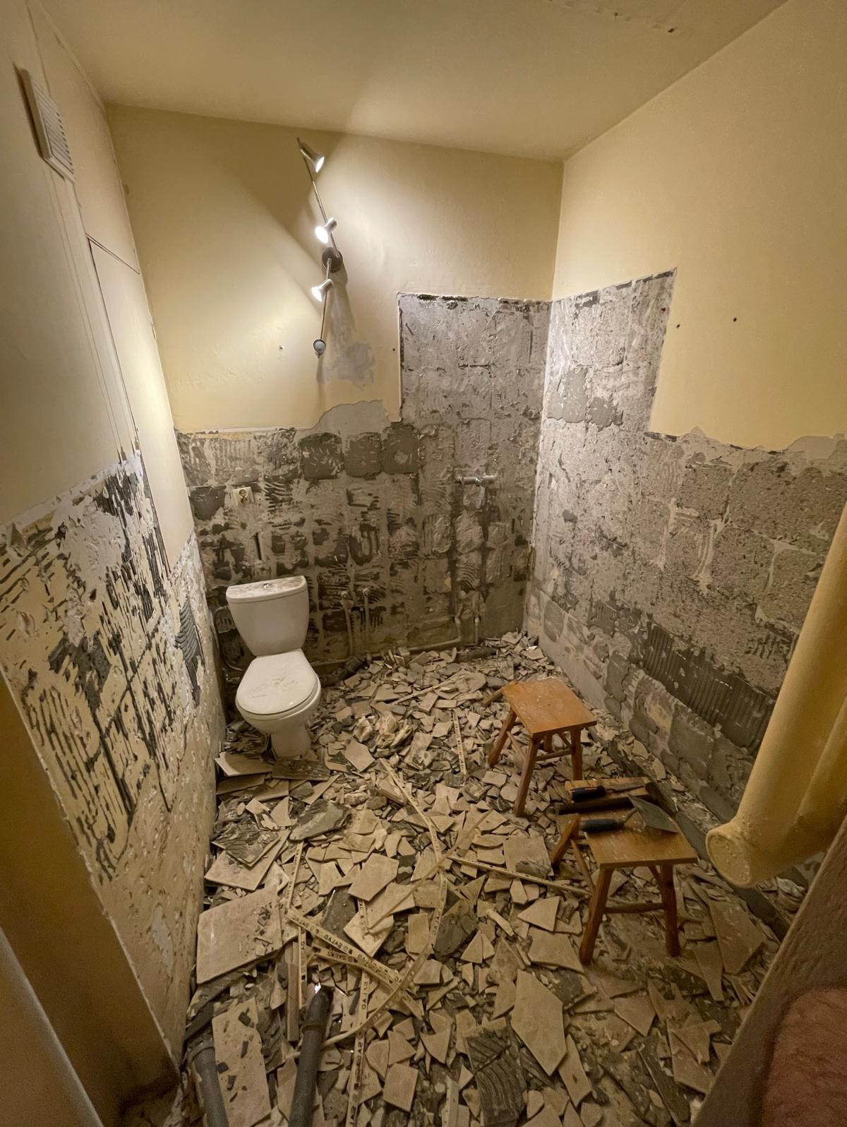 Demontaż płytek w łazience. Uwidocznione ściany z resztkami kleju, toaleta i gruz na podłodze. Dwa stołki glazurnicze z narzędziami. Etap przygotowania do remontu.