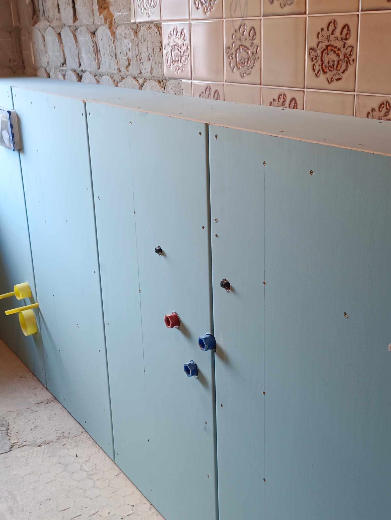 Stelaż z płyt gipsowo-kartonowych w łazience, z widocznymi przyłączami wodnymi (czerwone, niebieskie, żółte). W tle fragment starej ściany i płytek.