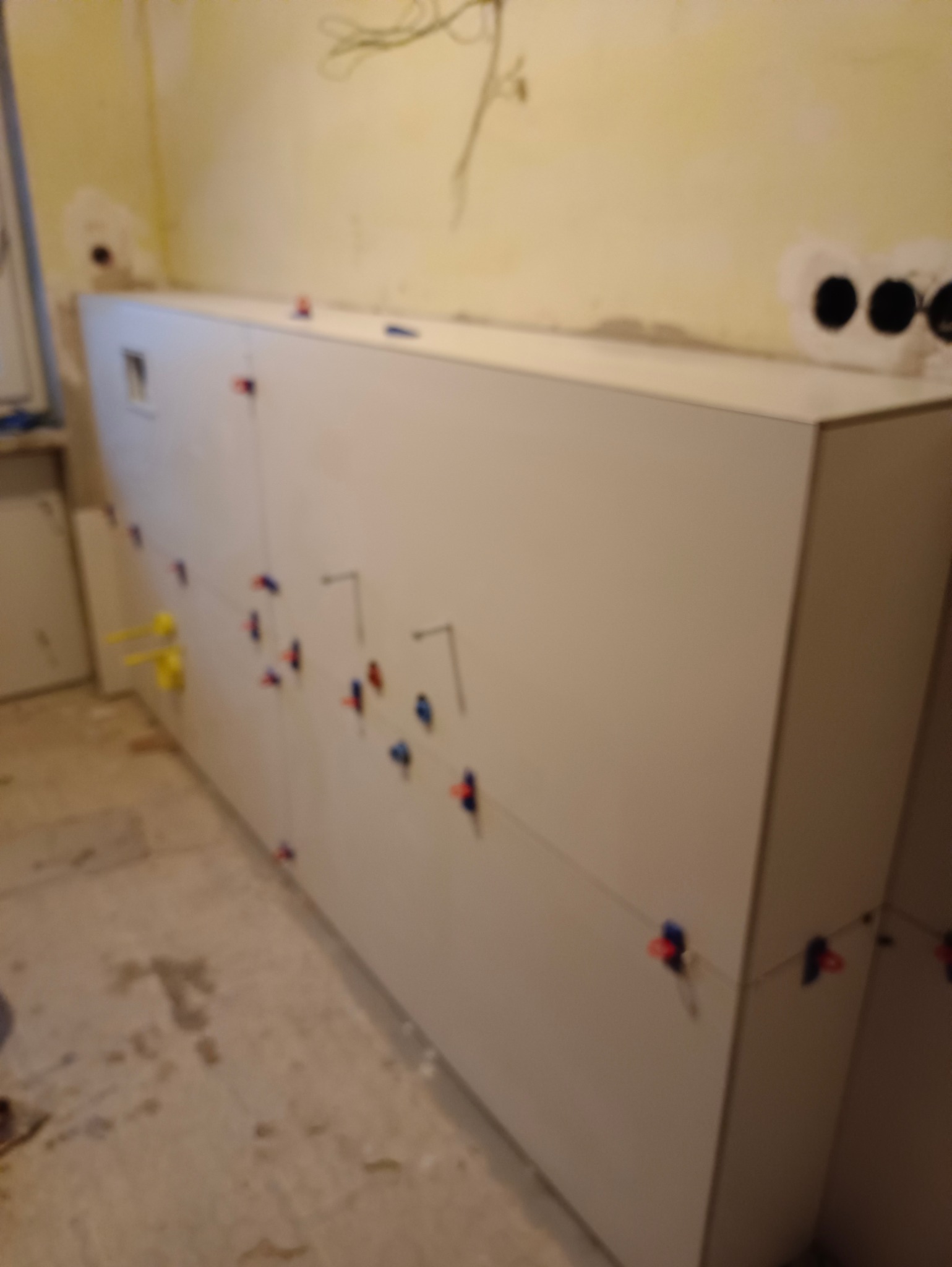 Białe płytki na ścianie w trakcie układania, z użyciem klipsów poziomujących. Widoczne przewody elektryczne i niedokończone ściany. Prace glazurnicze w łazience.