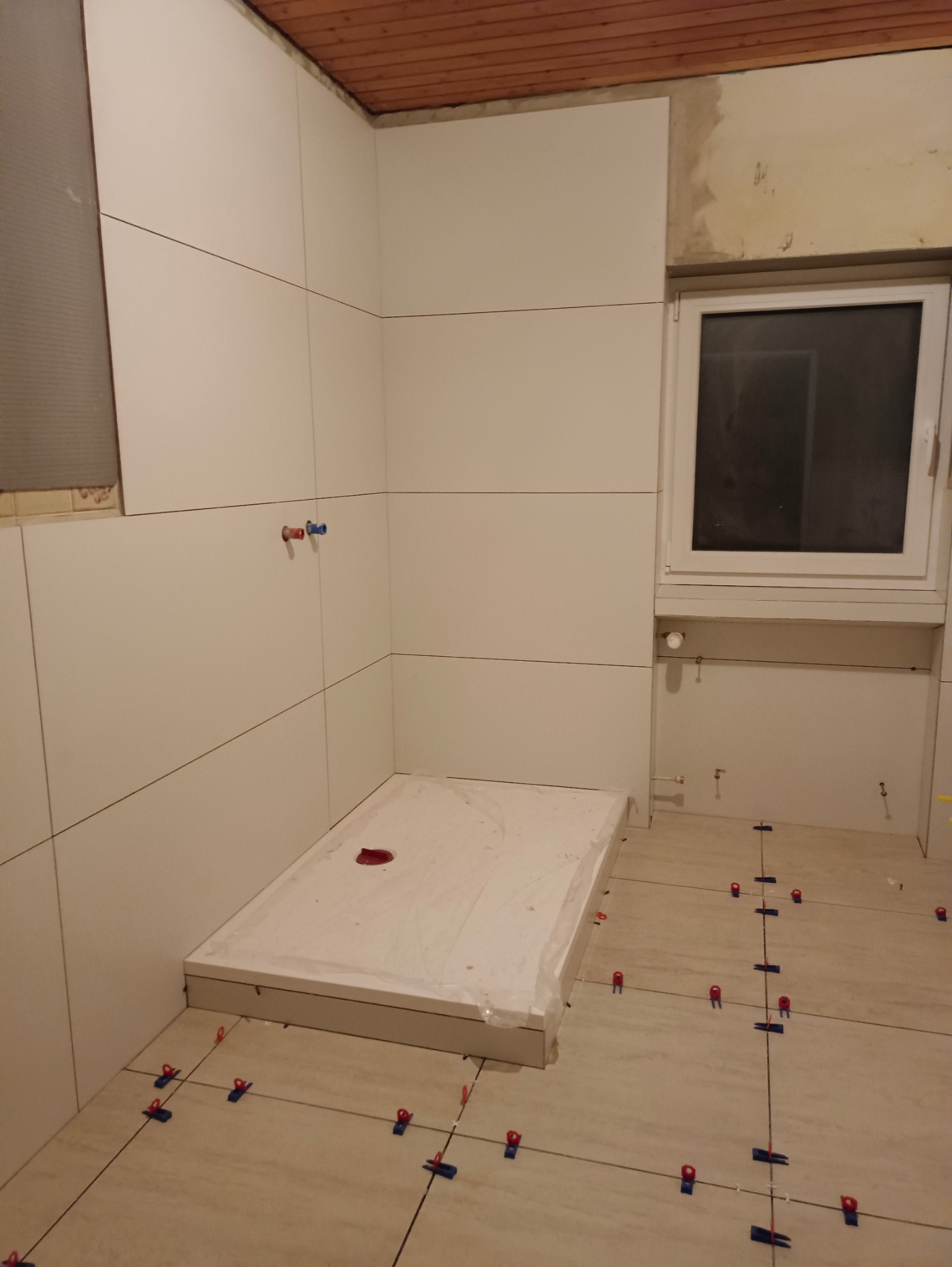 Białe płytki ścienne i podłogowe w trakcie układania w łazience. Widoczne krzyżyki dystansowe, odpływ liniowy i fragment drewnianej boazerii sufitowej.