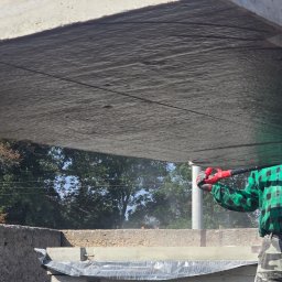 BIOTON SPÓŁKA Z OGRANICZONĄ ODPOWIEDZIALNOŚCIĄ - Montaż betonowej płyty drogowej za pomocą dźwigu przez pracownika w zielonej koszuli w kratę. Widoczne drzewa w tle i inne płyty.