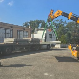 BIOTON SPÓŁKA Z OGRANICZONĄ ODPOWIEDZIALNOŚCIĄ - Żuraw przenosi betonową płytę drogową z ciężarówki na plac. Widoczne budynki w tle i błękitne niebo. Transport i rozładunek płyt.