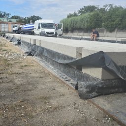 BIOTON SPÓŁKA Z OGRANICZONĄ ODPOWIEDZIALNOŚCIĄ - Ułożone płyty betonowe na placu budowy, zabezpieczone folią. W tle betoniarka i budynek. Mężczyzna siedzi na płycie, używając telefonu. Dzień, pochmurne niebo.