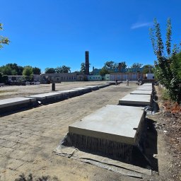 BIOTON SPÓŁKA Z OGRANICZONĄ ODPOWIEDZIALNOŚCIĄ - Układanie betonowych płyt drogowych na placu budowy w słoneczny dzień. Widoczny fragment placu z ułożonymi płytami, budynek w tle i komin. Prace w toku.