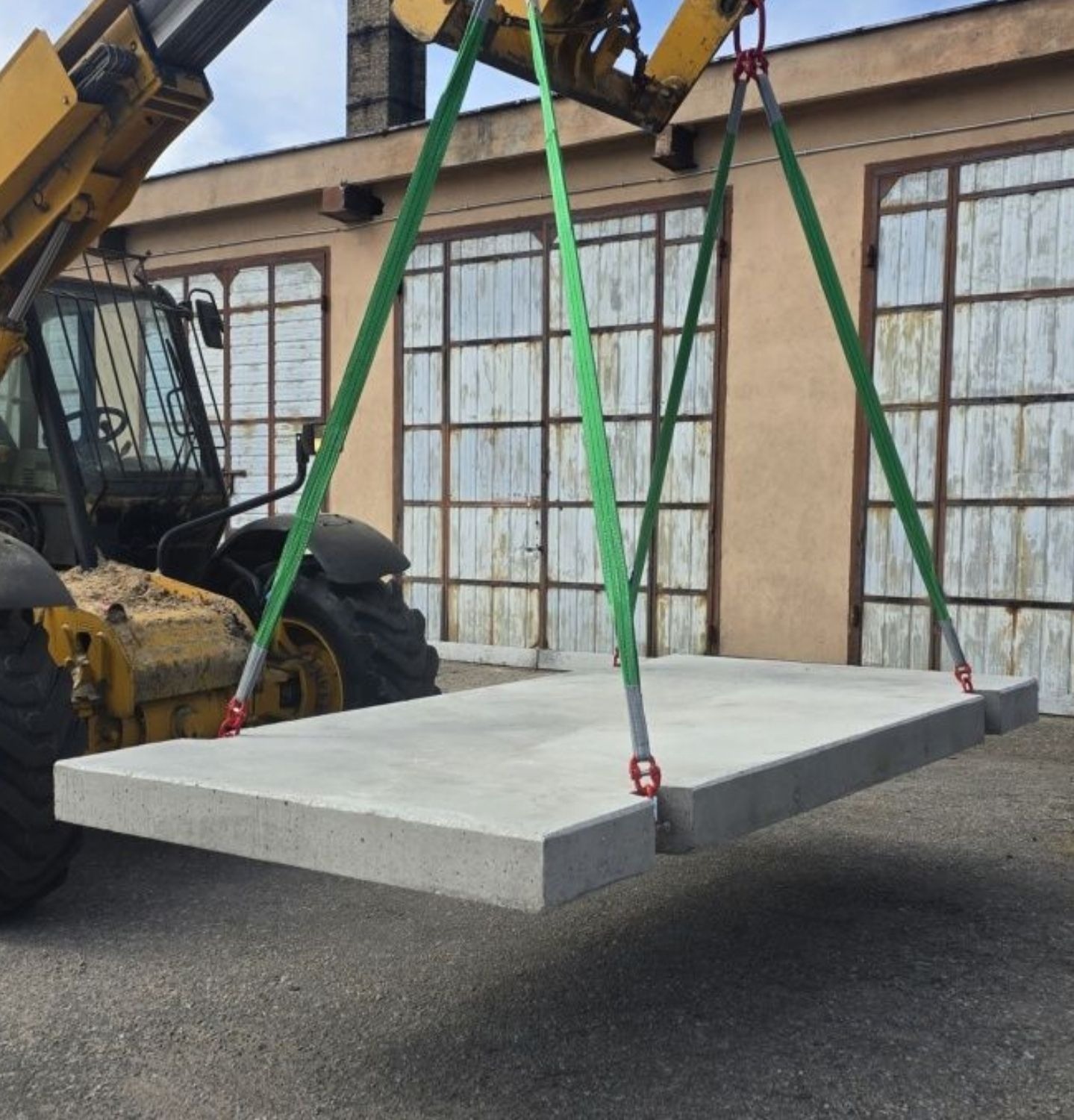 Żółty wózek widłowy podnosi betonową płytę drogową na tle budynku z dużymi oknami. Płyta zawieszona na zielonych pasach z czerwonymi elementami mocującymi.