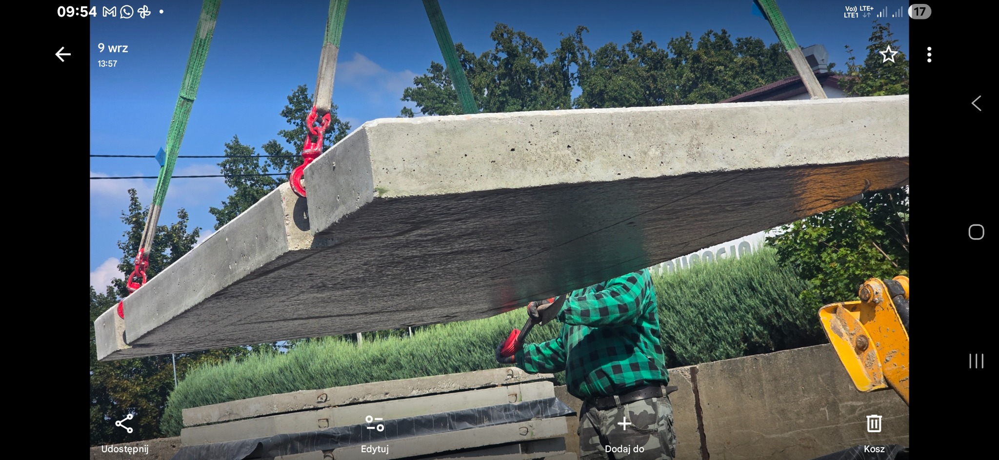 Montaż płyty drogowej za pomocą dźwigu. Widoczny pracownik w zielonej koszuli flanelowej i fragment koparki. Płyta betonowa zawieszona na pasach.