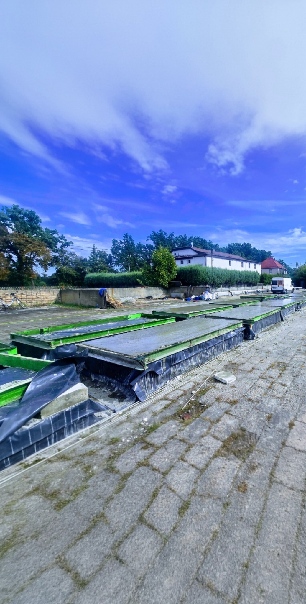 Formy do wylewania betonu na placu budowy, z widocznymi elementami konstrukcyjnymi i fragmentem nawierzchni z kostki. W tle budynek i błękitne niebo.